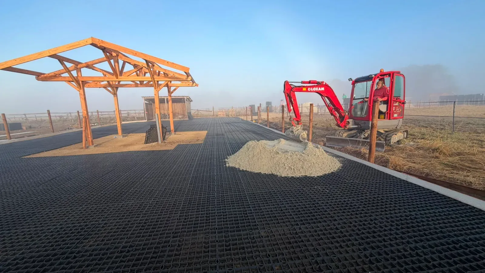 Mini-pelle et tas de sable sur l’aire stabilisée, abri en charpente bois à gauche